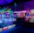 An indoor arcade filled with vibrant gaming machines lit with neon blue and pink lights. The scene includes various arcade games with flashing screens and colorful displays, and a large overhead structure resembling a green snake. The background shows illuminated signs and themed decor, contributing to an immersive entertainment atmosphere.